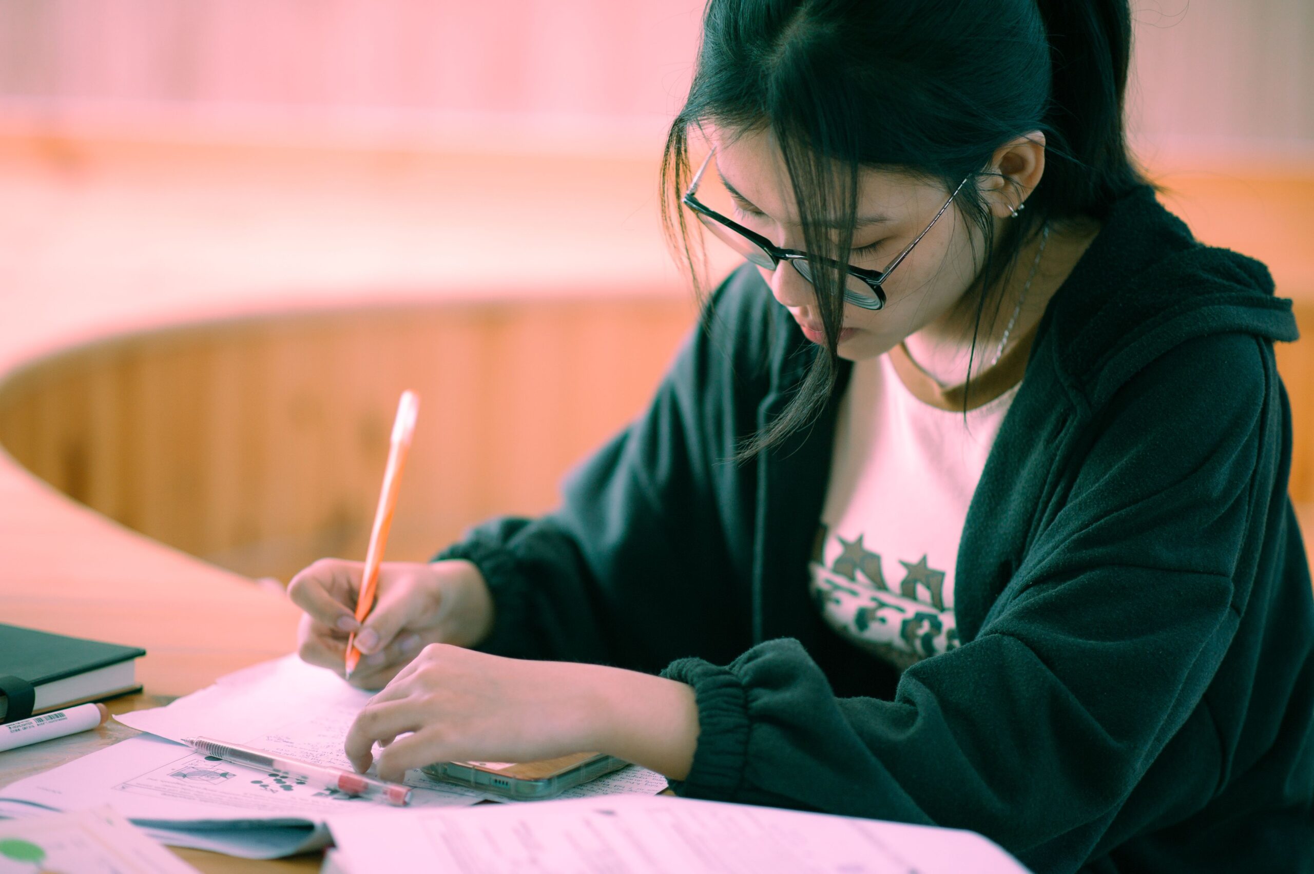 Asian woman wearing a hoodie and glasses, sitting at a table, immersed in writing.